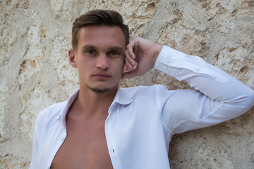 Portrait of the young handsome man in a white shirt standing near a stone wall