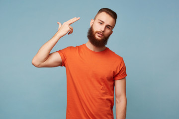 Portrait of young bearded man committing suicide with finger gun gesture. Portrait of despaired guy shooting himself making finger pistol sign against blue wall background. Human face expressions
