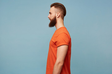 Portrait in profile of a guy with a fashionable hairstyle and a shaven head and a long thick beard in a red T-shirt standing sideways isolated on a blue background