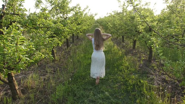 Carefree barefoot woman with flying curly blond hair running through spring apple orchard, rejoicing freedom in blooming nature. Joyful eco-friendly female in white dress relaxing in spring garden.