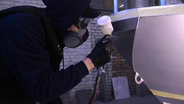 A professional guy (man) in the garage covers the yacht (transport) with a mini-jet (sprays) by applying paint (ceramics) to the surface using a protective suit dispenser and protective glasses.