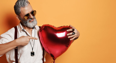 Senior bearded old man holds red heart ballon and pointing finger on it 