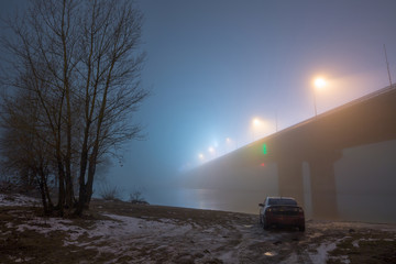A mysterious evening fog above the river in big city. Bridge in the mist, cold weather scenery. Soft, blurry, misty look. Colorful, mystic industrial cityscape. A car on the river bank.