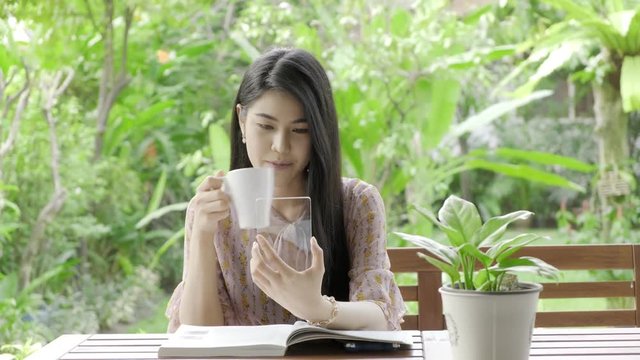Young beautiful Asian woman browsing phtos with high tech glass display in her home garden with green tree in background and drinking coffee. Future technology concept. For graphic overlay.