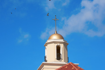 Beautiful church tower with golden cross and red tile roof against a bright blue sky with flying birds and fluffy clouds.