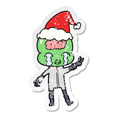 distressed sticker cartoon of a big brain alien crying and giving peace sign wearing santa hat