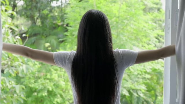 Slow motion of young beautiful asian woman open bedroom curtain to see green trees background. Relaxing home concept.