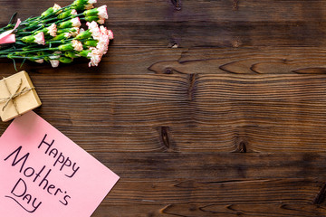 Happy Mother's Day concept. Hand lettering near bouquet of pink carnation and gift box on dark wooden background top view copy space