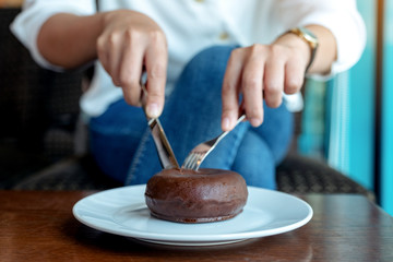 Fototapeta na wymiar Closeup image of woman's hands cutting a piece of chocolate donut by knife and fork for breakfast on wooden table