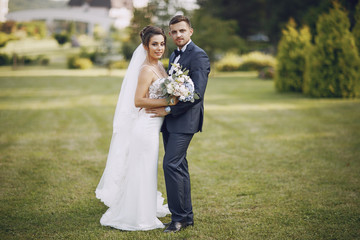 A young and beautiful bride and her husband is standing in a summer park with bouquet of flowers
