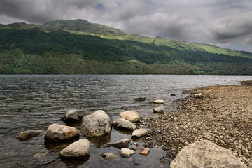 Fototapeta premium Rocky shore of Loch Lommond freshwater lake dividing lowlands from Scottish Highlands and Ben Lamond mountain Scotland UK