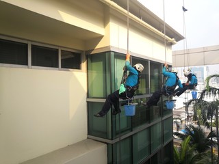 Rope access cleaning worker wearing safety harness hard hat working at height descending on rope performing washes a hospital complex glass wall.