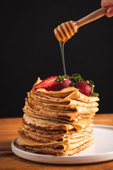Pouring honey over stack of blini, crepes, thin pancakes. On black background with copy space for text. Tasty breakfast food, dessert, traditional Russian food for Maslenitsa
