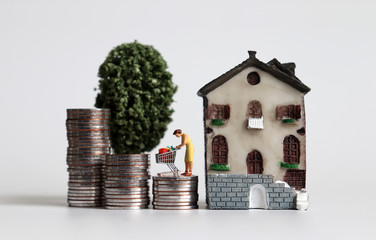 A miniature woman standing with a shopping cart on three pile of stepped coins next to a miniature house with a miniature tree.