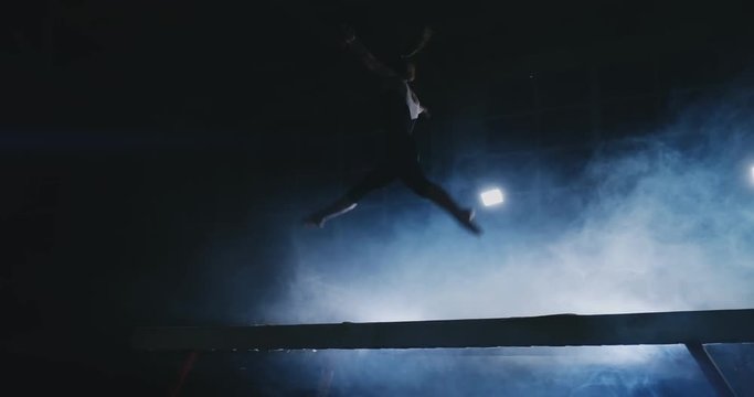 The girl is a professional athlete performs gymnastic acrobatic trick on a beam in backlight and slow motion in sports gymnastic clothing. Smoke and blue. Jump and spin on the balance beam
