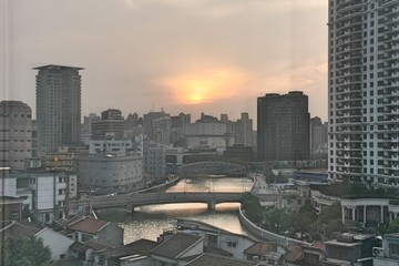 Cityscape in Shanghai with skyscrapers, high rise and low rise buildings, and houses with bridge and river at dusk in front of sunset in cloudy sky