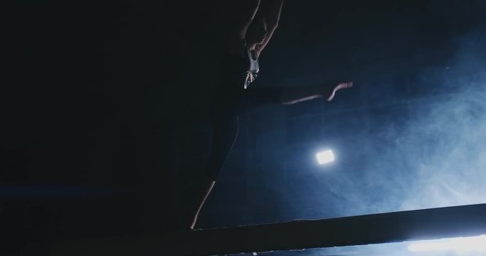 Side view of a female gymnast doing split handstand on balance beam against black background.