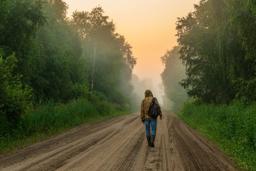 a man walking on a forest road with a backpack. misty morning in the green forest. walking along a sandy road surrounded by trees