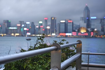 Fototapeta na wymiar Metal handrail with blurred bokeh background of Hong Kong Island skyline cityscape and green leaves of bush under cloudy gray sky