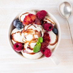 Balls of vanilla ice cream in mug with raspberry and blueberries, chocolate syrup  topping. Top view summer dessert, white, bright background, macro