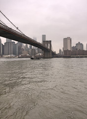 Manhattan skyline and Brooklyn bridge with river in foreground on cloudy, rainy day with gray sky