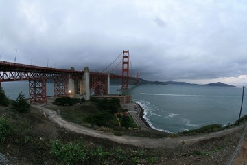 Golden gate bridge with ocean in foreground and hills and mountains in background under gray cloudy sky on rainy day