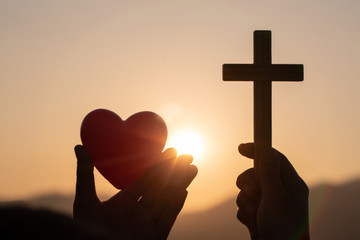 Silhouette of woman hands praying with cross and holding a red heart ball  in nature sunrise...