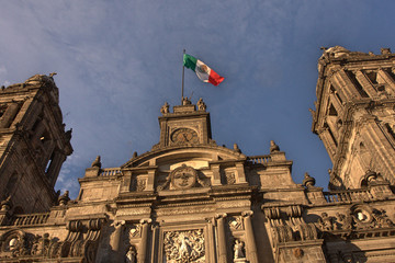 Facade of sandstone church building as seen from below with cross symbols and bell towers and Mexican flag flapping in wind under bright blue sky