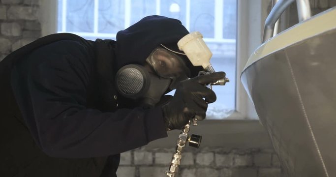 A professional guy (man) in the garage covers the yacht (transport) with a mini-jet (sprays) by applying paint (ceramics) to the surface using a protective suit dispenser and protective glasses.
