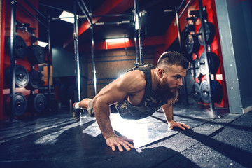 Strong hipster bodybuilder doing push ups on the gym floor with weight vest on. Gym interior, night workout concept. - obrazy, fototapety, plakaty