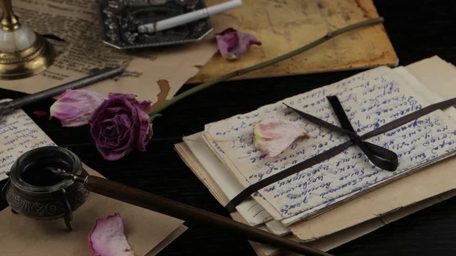 Burning candle in a candlestick on the background of the dried rose, cigarette in an ashtray, an old letter, envelopes and inkwell with a pen. Memory, family archive