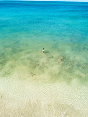 Aerial view of a girl in red swimming suit walking into the Andaman sea of green color. There is blue sky and white sand beach. Koh Lanta, Thailand, Asia.