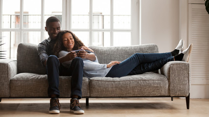 Happy mixed race couple relax on couch using smartphone