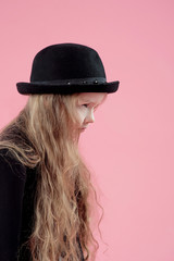 funny cute serious little girl with long blonde hair in black blazer and stylish black hat on pink background