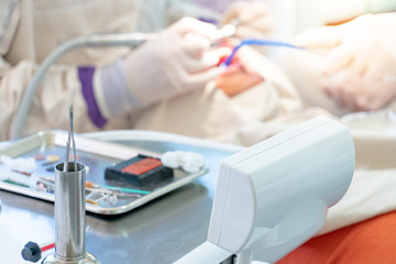 Dental instruments and medical supplies on metal tray in dental clinic with blurred dentist and assistant curing patient teeth in the background.