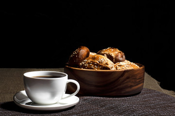 A cup with a hot drink and a wooden plate with pies stand on a linen tablecloth against a black background.