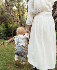 Mom in a white dress walks and holds a curly son by the hand in a green park in the summer.