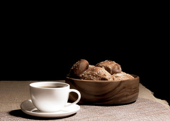 A cup with a hot drink and a wooden plate with pies stand on a linen tablecloth against a black background.