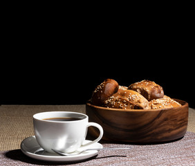 A cup with a hot drink and a wooden plate with pies stand on a linen tablecloth against a black background.