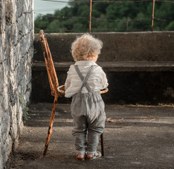 A little boy with curly white hair in gray pants and a white shirt stands near an old rusty chair in front of a stone wall