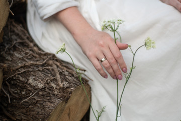 A girl in a white dress sits on a wooden log and touches a flower. On hand a wedding ring. Country Wedding,