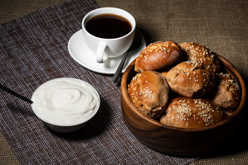 Pies in a wooden plate, a cup with a hot drink and a plate with sour cream stand on a table covered with a linen tablecloth.
