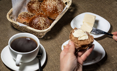 Female hand puts butter on a sliced sesame bun. A cup of hot drink stands nearby.
