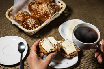 Female hand puts butter on a sliced sesame bun. A cup of hot drink stands nearby.