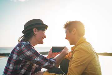 Happy lesbian couple watching on mobile phone next the beach at sunset - Young homosexual women having fun with new trends technology - Lgbt, homosexuality, tech and relationship concept