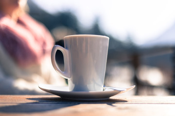 Cup of coffee on the table of a café, outdoors in the sunny afternoon. Woman in the blurry background.