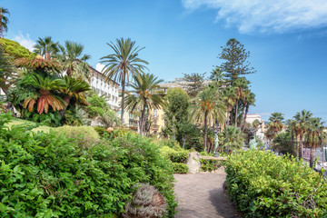 Fototapeta na wymiar Beautiful plants and blooming flowers with two statues of handsome women holding a torch in a garden along the coastal road from San Remo