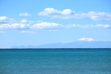 Obraz premium High mountains silhouettes with white snow cap on top far away on horizon in light haze after the sea under blue sky with white cumulus clouds