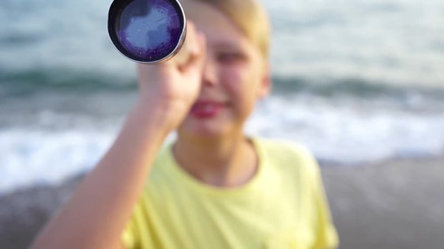 Closeup view of cute happy young kid standing at summer beach and looking through vintage old hendheld telescope at something interesting far away in distance. Real time 4k video footage.