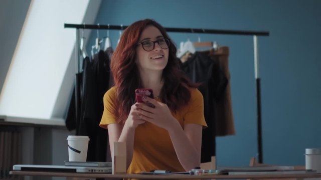A stylish young woman in a bright T-shirt is sitting at the table, looking at the smartphone screen is reflected in the glasses. Her colleague moves the coat hanger and outweighs the dresses.